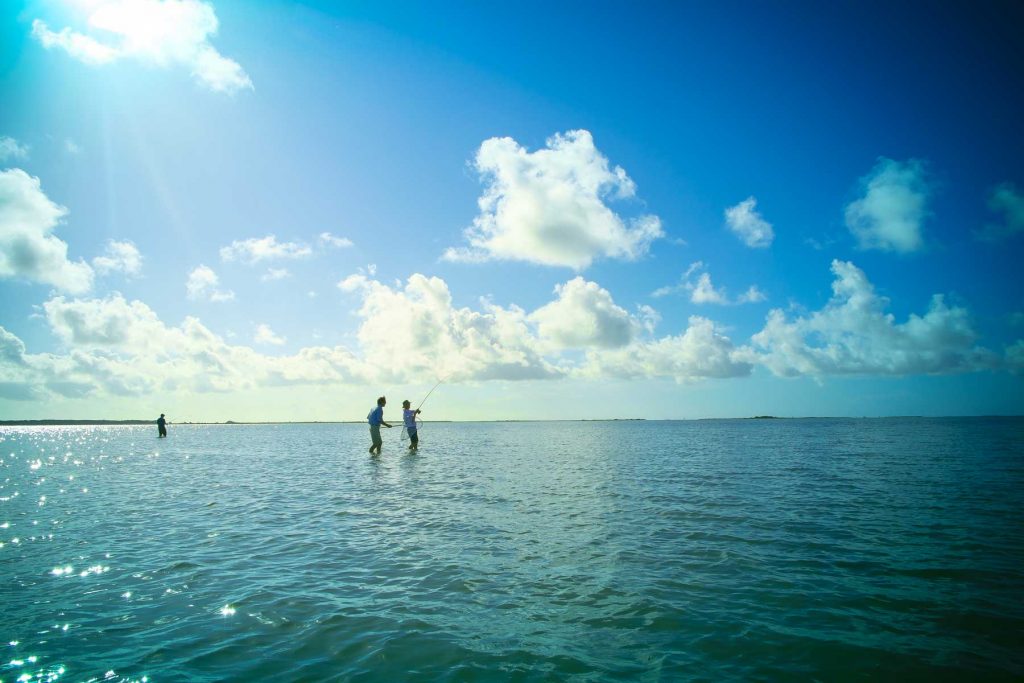 Anglers fly fishing in shallow turquoise water under a bright sunny sky.