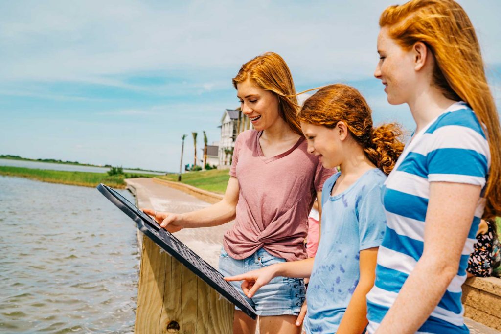 Three redhead girls examining an informational sign on a waterfront boardwalk.