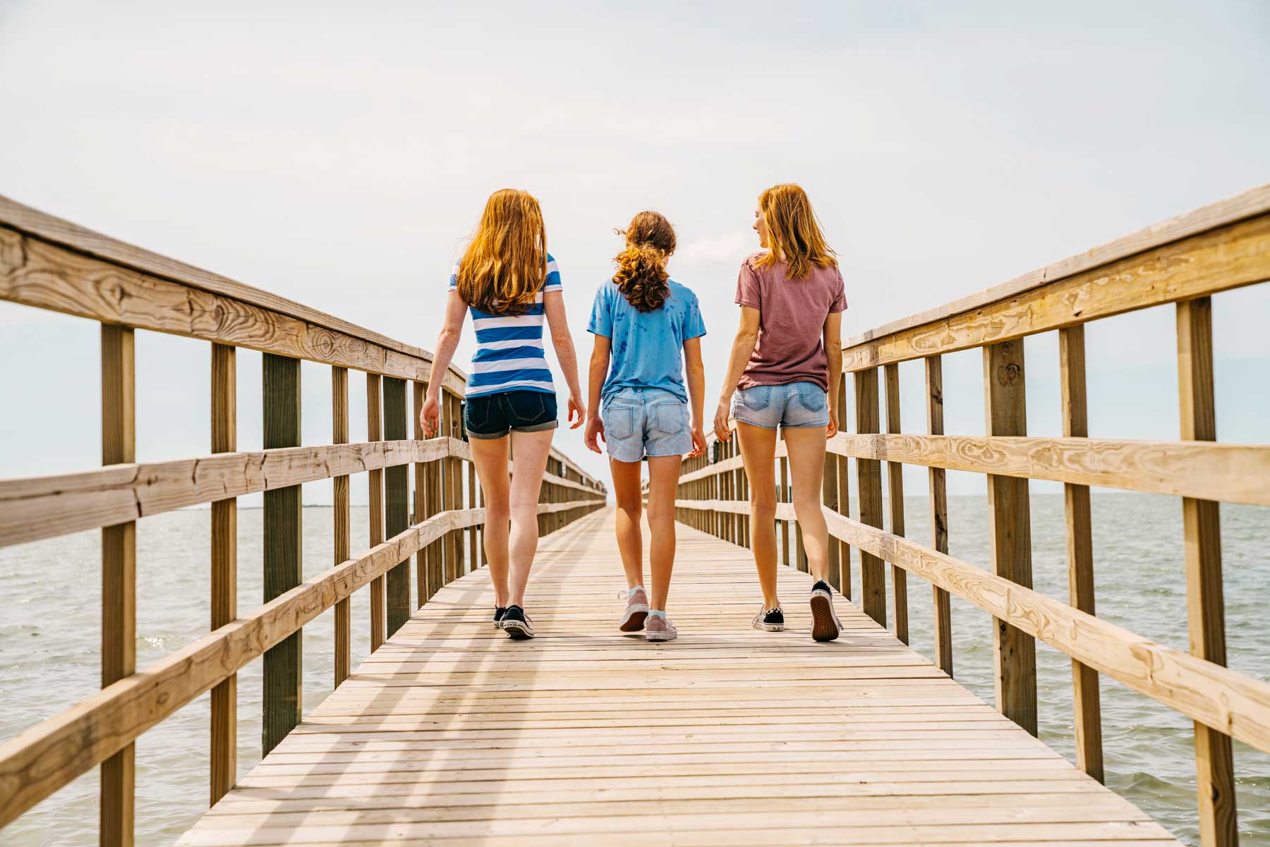 Three young women walking away on a long wooden pier over the water.