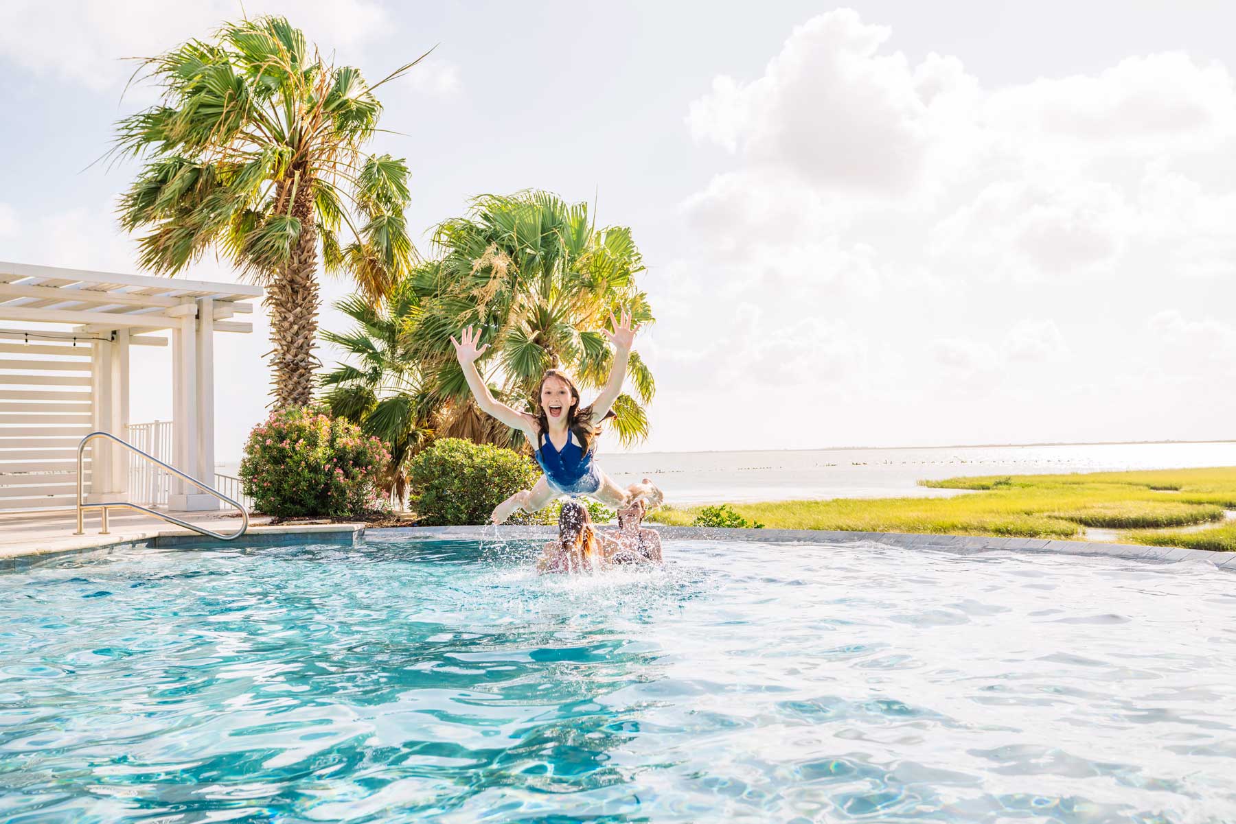 Girl tossed into the air above an outdoor swimming pool with palm trees.