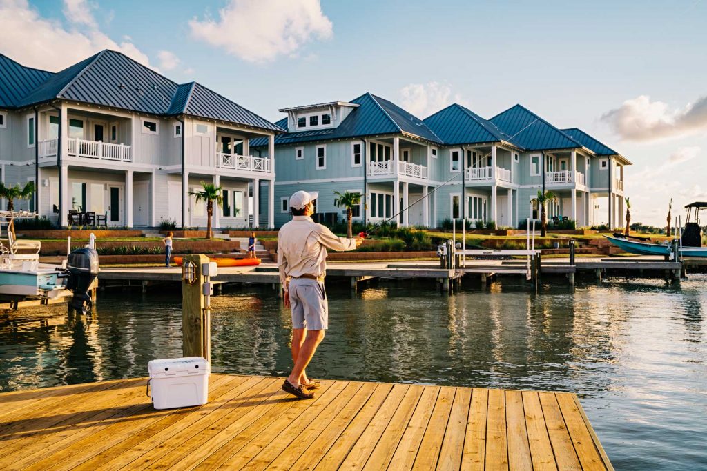 Man fishing from a wooden dock in front of modern coastal waterfront homes.