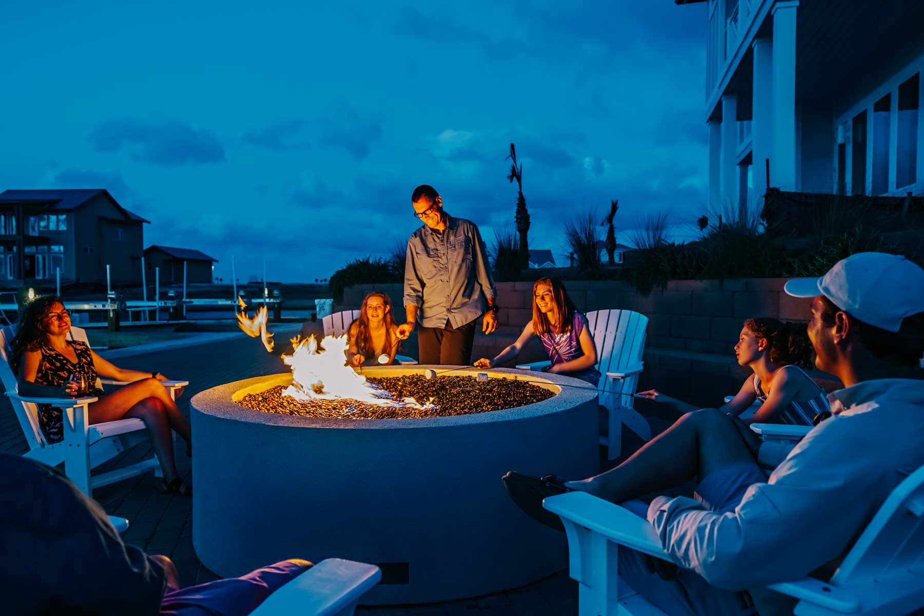 People roasting marshmallows over a large circular fire pit on a waterfront patio at dusk.