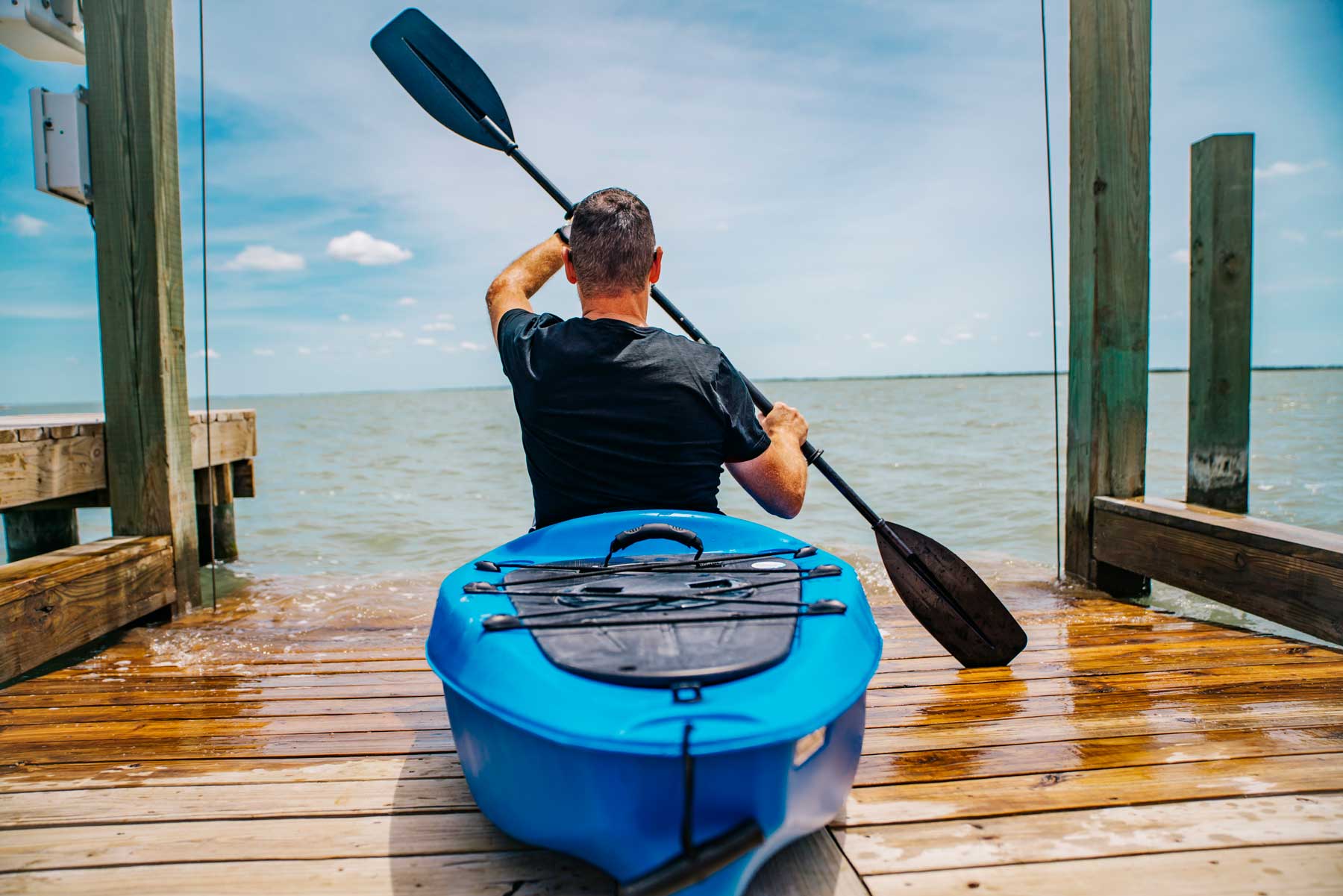 Man in a blue kayak on a wooden dock holding a paddle over water.