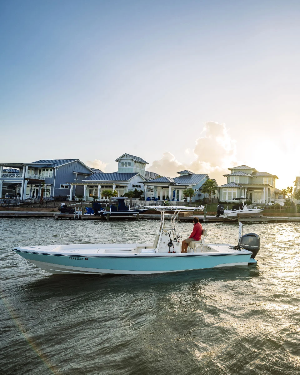 Man piloting a light blue boat past waterfront houses during golden hour.
