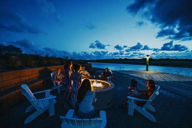 Group gathering around a fire pit on a waterfront patio at dusk.
