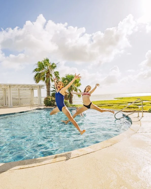 Two girls jumping into a swimming pool with palm trees and coastal views.